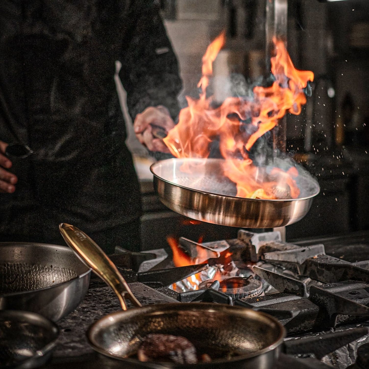 Person cooking over a flaming stove with multiple pans on a stovetop.