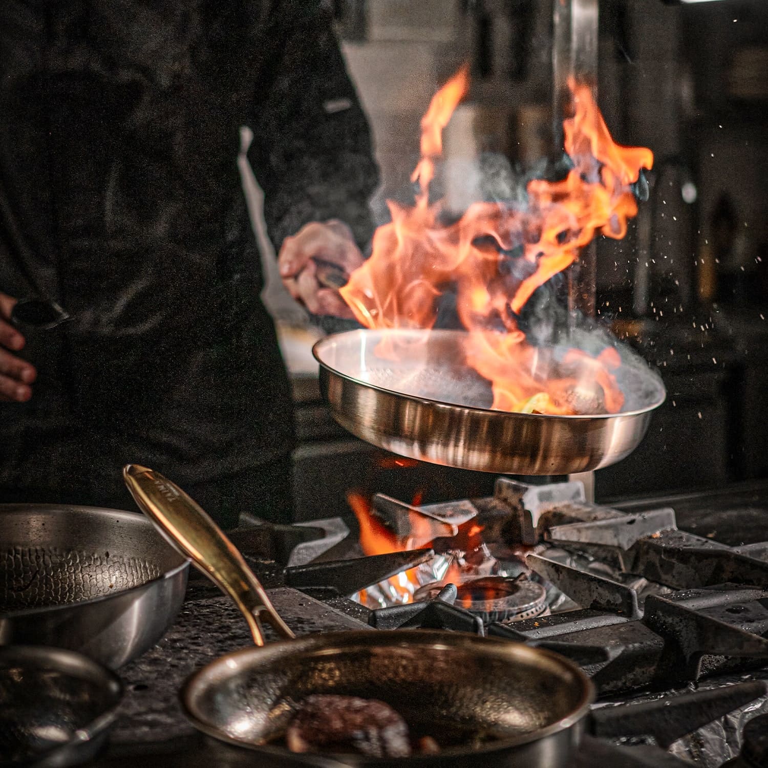 Person cooking over a flaming stove with multiple Titanium pans on a stovetop.